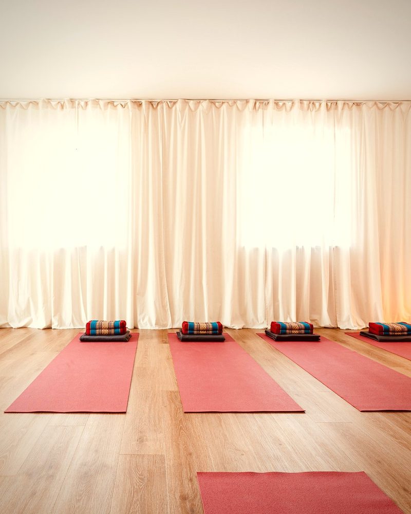 Der große Yogaraum mit lichtdurchfluteten Vorhängen und roten Matten